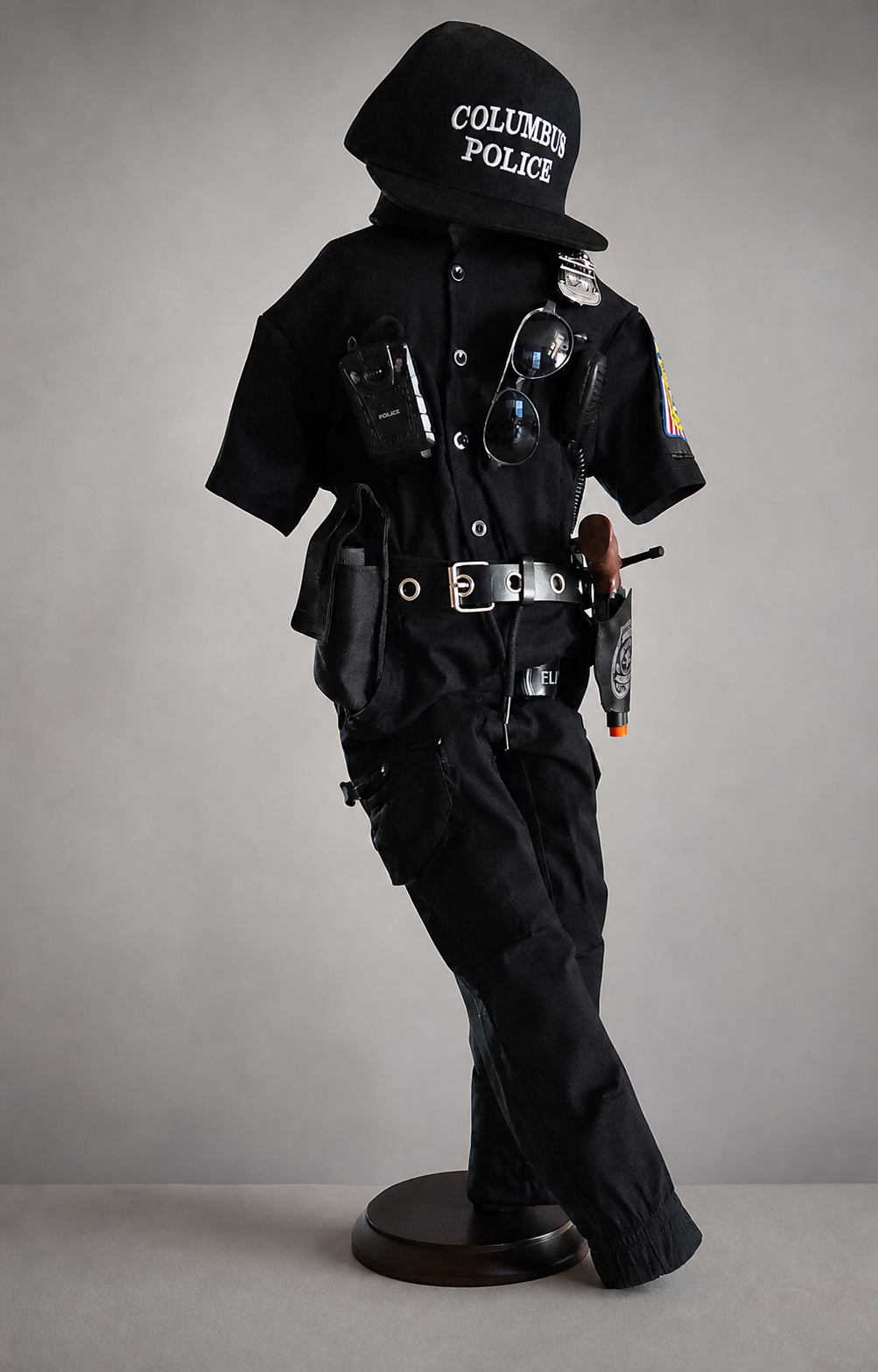 Child-size black Columbus Police uniform on a stand: cap, short-sleeve shirt with patch and badge, duty belt with toy radio and holster, and cargo pants